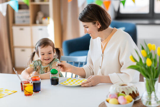 holidays, family and people concept - happy mother and little baby daughter dyeing easter eggs with colors at home