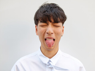 Portrait of Chinese young man with black hair in white shirt posing against white background, making face eyes closed with tongue out, dislike expression, front view.