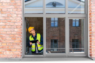 architecture, construction business and building concept - male architect in helmet and safety west with tablet pc computer in window