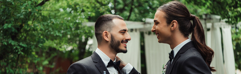 side view of cheerful gay newlyweds in suits looking at each other, banner. - Powered by Adobe