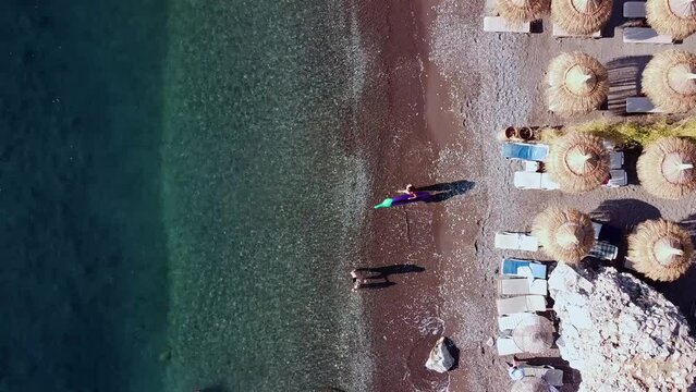 Woman with air mattress for swimming going to sea water. Funny eggplant inflatable mattress. Aerial top view on people swimming in the sea and people relaxing on the beach.