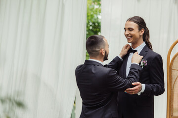 gay man adjusting bow tie on suit of happy groom in formal wear.