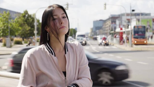 A young beautiful Caucasian woman looks around in thought in a street in an urban area - busy roads in the background