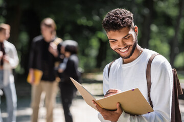 Smiling african american student with backpack writing on notebook outdoors.