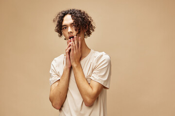 Confused awesome tanned curly man in basic t-shirt touches cheeks posing isolated on over beige pastel background. Fashion New Collection offer. People and Emotions concept. Free place for ad