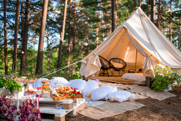 Beautiful white decor in boho style. Picnic in the nature, table, carpets, wigwam, tent, pillows in...