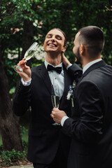 positive gay newlyweds in suits holding glasses with champagne on wedding day.