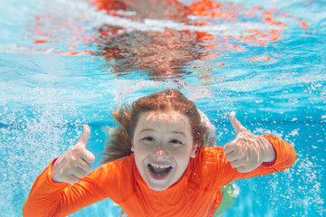 Excited kid boy with thumbs up swim and dive underwater, kid with fun in pool under water. Active healthy lifestyle, water sport activity and swimming lessons on summer vacation with child.