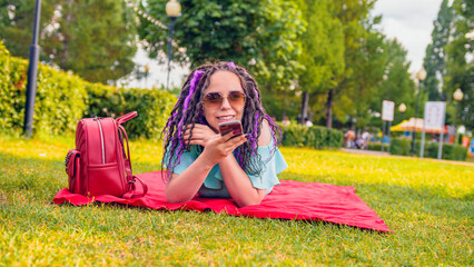 Young woman lying on a blanket records a voice message using a smartphone in the park. Happy girl...