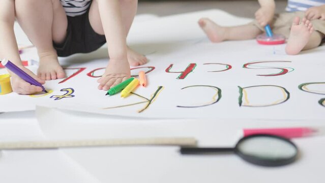 unrecognizable hands and barefoot legs of children kids, drawing back to school words on white paper lying on floor in living room home indoors. concept of first day at elementary school. 