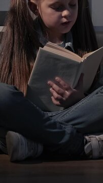 Vertical video. Kid reading hobby. Intelligent child. Night fairy tale. Curious smart cute little girl with open book sitting on floor cross-legged.