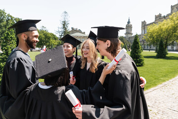 Smiling interracial bachelors in gowns holding diplomas and hugging in park.