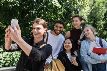 Smiling student taking selfie on smartphone near interracial friends in park.