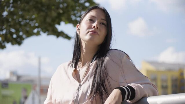 A young beautiful Caucasian woman looks into distance in thought as she listens to music with earphones on in an urban area