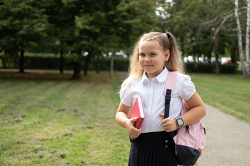 blonde smiling schoolgirl in school uniform holding notebook with pink backpack back to school outdoor