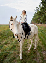 Little smiling girl riding  white blue eyed horse in the autumn evening forest 