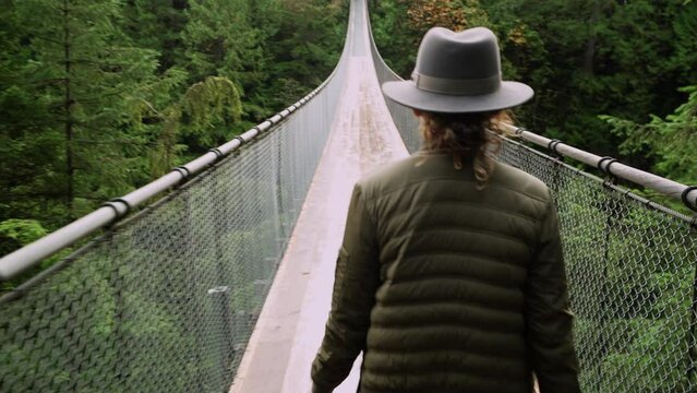 Female traveler wearing a hat, walking along suspension bridge in lush rainforest setting in North Vancouver, British Columbia, Canada. 