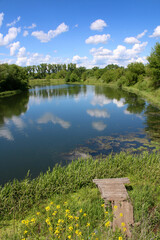 Fototapeta premium European landscape. A small calm river and a wooden fishing pier on the shore. Clouds reflect in the river.