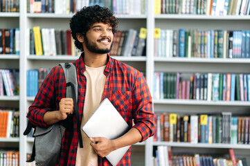 Positive confident indian or arabian male student of university, in stylish casual wear, with backpack and laptop, stands in a library against the background of bookshelves, looks away, smiles, dreams