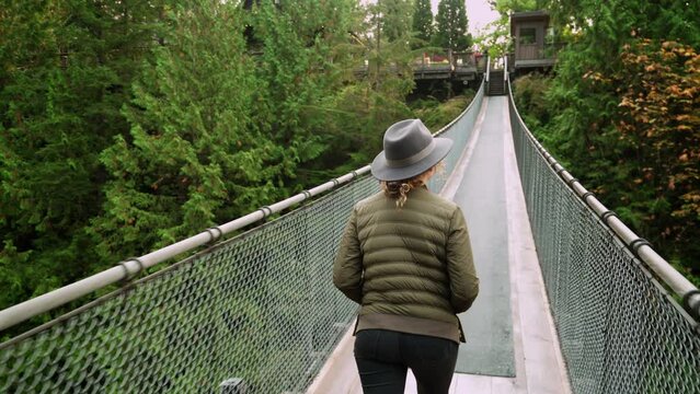 Tourist woman wearing a hat, walking along suspension bridge in lush rainforest setting in North Vancouver, British Columbia, Canada. 