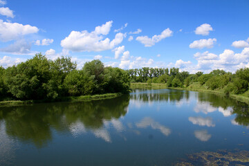 Fototapeta na wymiar European landscape. A small calm river and densely forested banks. Clouds reflect in the river.