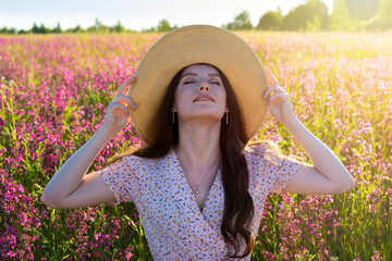 A beautiful young woman in a hat stands on a flowering meadow in the sun. Enjoys summer, sunlight, warm weather.