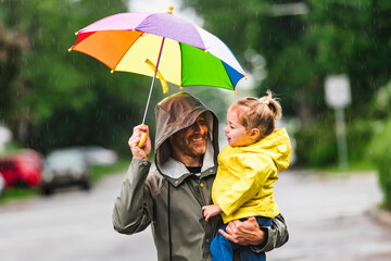 Happy funny father holding multicolored umbrella under the summer raining day with the child girl - obrazy, fototapety, plakaty