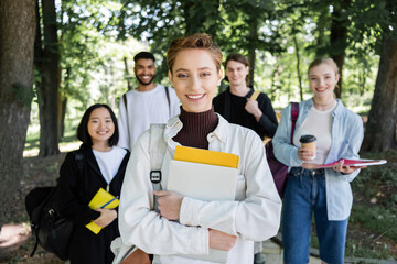 Smiling student with laptop and notebook looking at camera near blurred interracial friends in park.