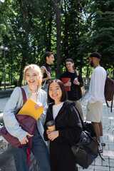 Interracial students with notebook and coffee to go smiling at camera near blurred friends in park.