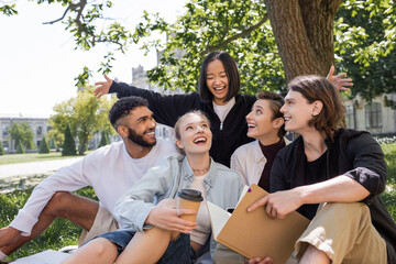 Excited asian student looking at cheerful interracial friends on grass in park.