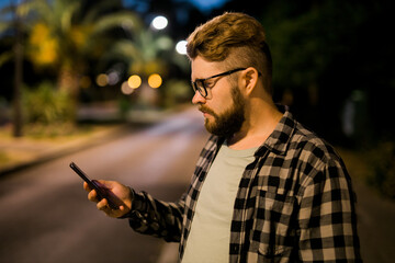Bearded man wearing eyeglasses is holding scrolling texting in his cellphone at night street. Guy calls for taxi in an app in evening city - copy space and place for advertising