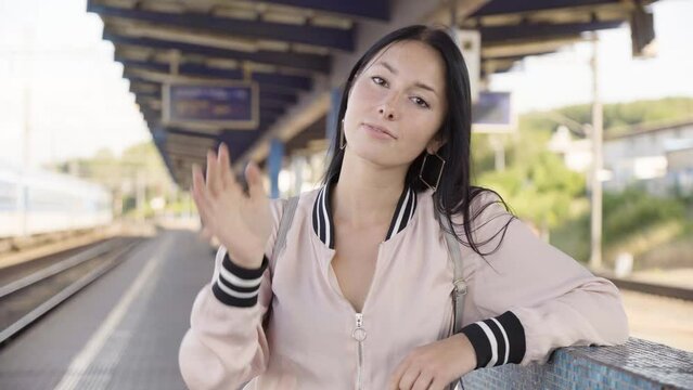 A young beautiful Caucasian woman waves at the camera with a smile at a train station