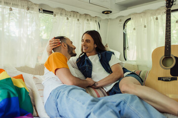smiling gay man with tattoo looking at bearded boyfriend on bed near lgbt flag and acoustic guitar...