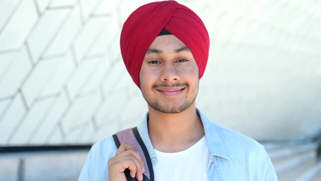 Modern male Indian student with backpack wearing red national headwrap standing outdoors, hindu guy in traditional headdres looking at the camera with pleasant smile