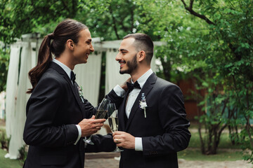 side view of cheerful gay newlyweds in suits holding glasses with champagne on wedding day.