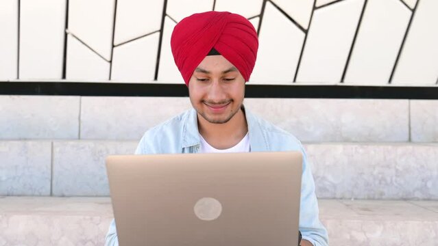 Cheerful Indian man wearing headwrap sitting on the steps outdoors and using laptop. Inspired glad male entrepreneur typing on keyboard and looks at the screen with pleasure smile