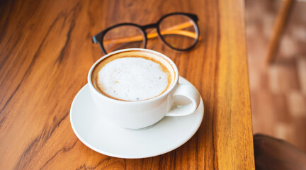 Cup of cappuccino coffee with latte and glasses of a woman on a wooden bar counter in a sunlit cafe in the morning.