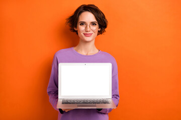 Photo of charming lady hold demonstrate empty space netbook display isolated on orange color...