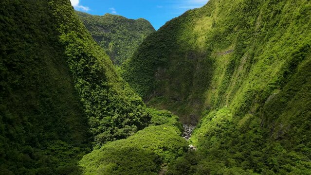 Aerial view of a ravine on the Reunion Island, sunlight break through the clouds, the mountain is covered in green vegetation. At the bottom there is a river. The sky is blue.