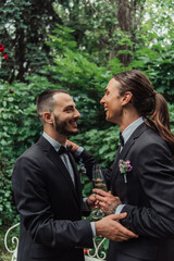 side view of happy gay newlyweds in suits holding glasses with champagne on wedding day.