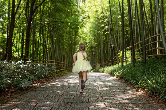 Cute Little Girl In Yellow Tutu Dress Running In Bamboo Forest In China. Kid With Blonde Hair From Behind Outdoor