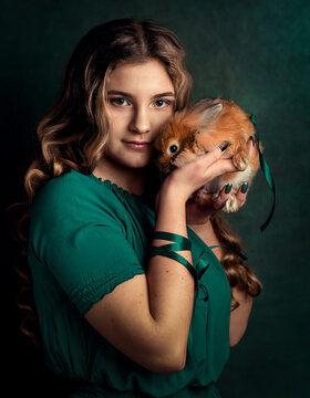 A brunette with long curly hair in a green cyan dress holds a red rabbit in her hands.
The photo is made in retro style.