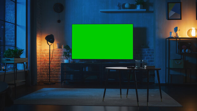 Stylish Loft Apartment Interior with TV Set with Green Screen Mock Up Display Standing on Television Stand. Empty Living Room at Home with Chroma Key Placeholder on Monitor. Night Shot.