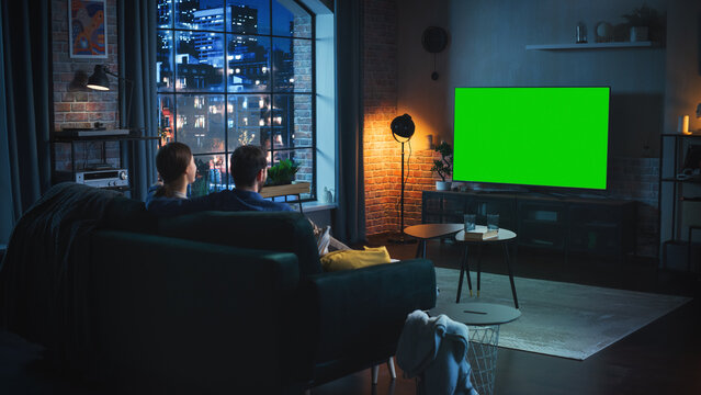 Beautiful Couple Spending Time at Home, Sitting on a Couch and Watching TV with Green Screen Mock Up Display in Their Stylish Loft Apartment. Man and Woman Streaming Movie or Show and Eating Popcorn.