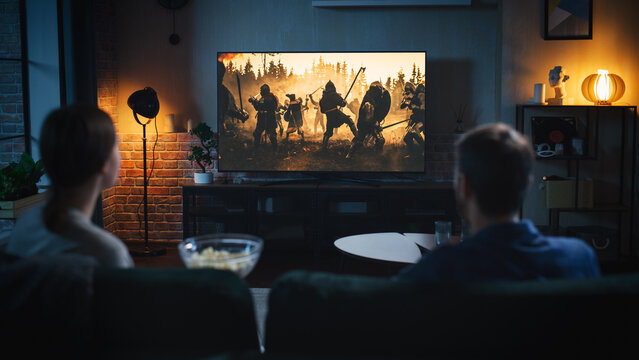 Authentic Couple Spending Time at Home, Sitting on a Couch and Watching Latest Blockbuster on Flat Screen Television Set. Man and Woman Streaming Movie or Show Using Home Cinema System.