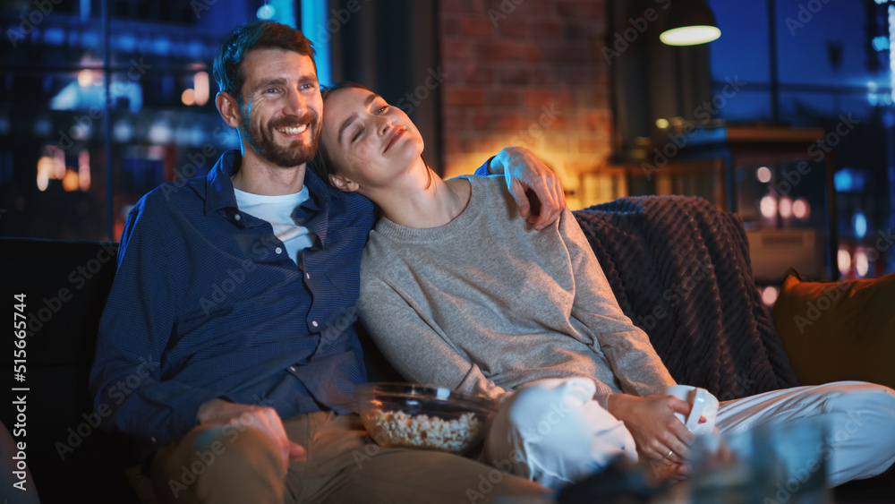 Wall mural Portrait of Beautiful Couple Spending Time at Home, Sitting on a Couch, Hugging and Watching Exciting TV Show in Their Stylish Loft Apartment. Man and Woman Streaming Comedy Movie and Have Good Time.