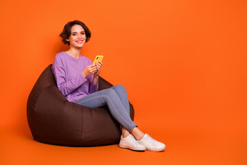 Full body portrait of cheerful nice lady sitting comfort bag use telephone isolated on orange color background
