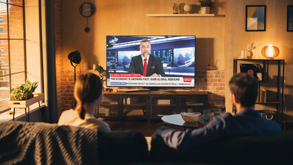 Beautiful Couple Watching News On Television Set in Cozy Loft Apartment. Young Man and Woman are...