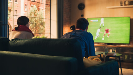Couple of Soccer Fans Relax on a Couch, Watch a Sports Match at Home in Stylish Loft Apartment. Relaxed young Man and Woman Cheer for Their Favorite Football Club and Enjoying the Weekend.
