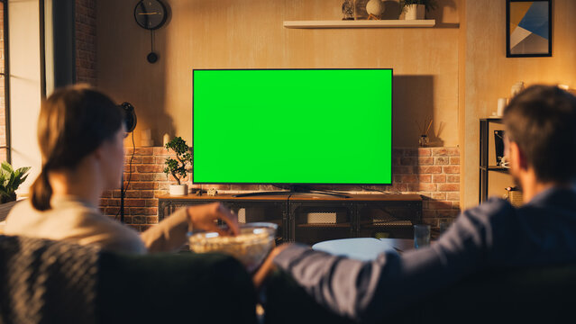Authentic Couple Spending Time at Home, Sitting on a Couch and Watching TV with Green Screen Mock Up Display in Their Stylish Loft Apartment. Man and Woman Streaming Movie or Show. Shot From Back.